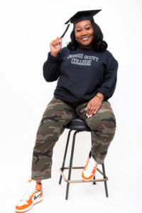 Lexie is sitting on a stool while wearing a black Agnes Scott College crewneck, camp pants, and orange sneakers. The picture has a white background